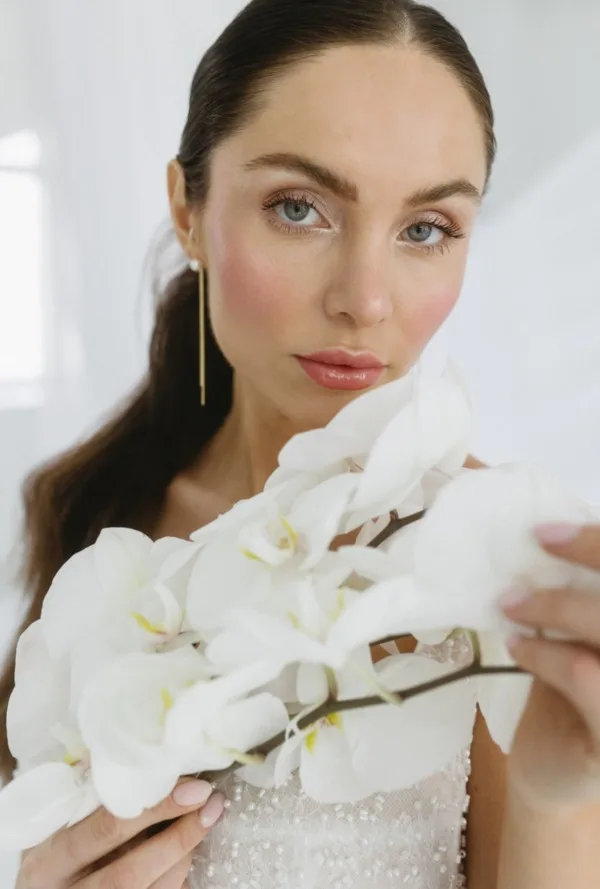 Dunkelhaarige Braut schaut direkt in die Kamera. Sie hat Orchideen in der Hand und trägt Brautohrringe
